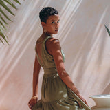 A woman with short hair poses in a flowing sage green Boho Bridesmaid Dress by AYA Sacred Wear, looking over her shoulder. Sunlight filters through palm fronds, casting shadows on a peach-colored wall. She wears gold earrings and bracelets, creating an elegant, tropical ambiance.