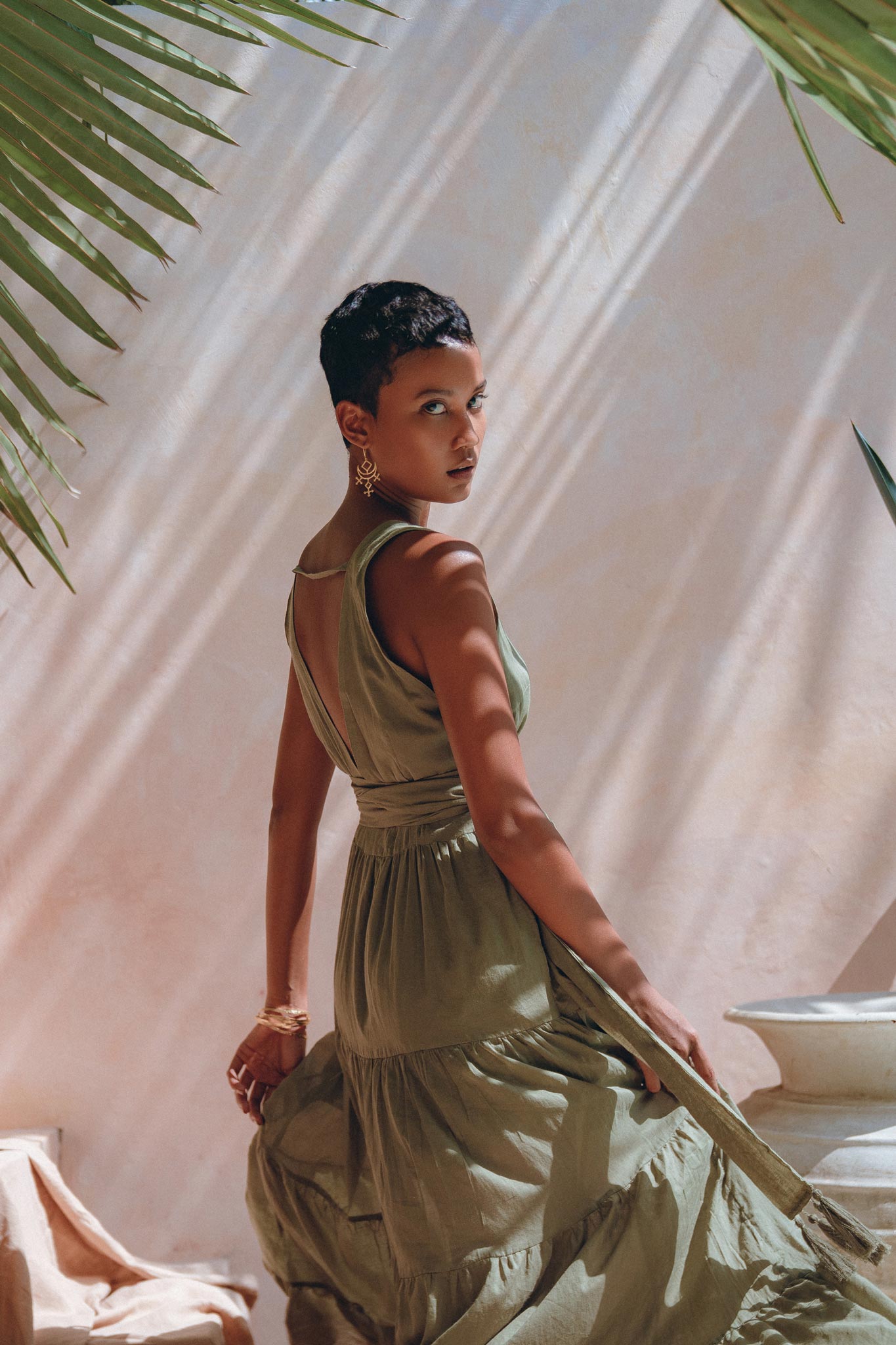A woman with short hair poses in a flowing sage green Boho Bridesmaid Dress by AYA Sacred Wear, looking over her shoulder. Sunlight filters through palm fronds, casting shadows on a peach-colored wall. She wears gold earrings and bracelets, creating an elegant, tropical ambiance.