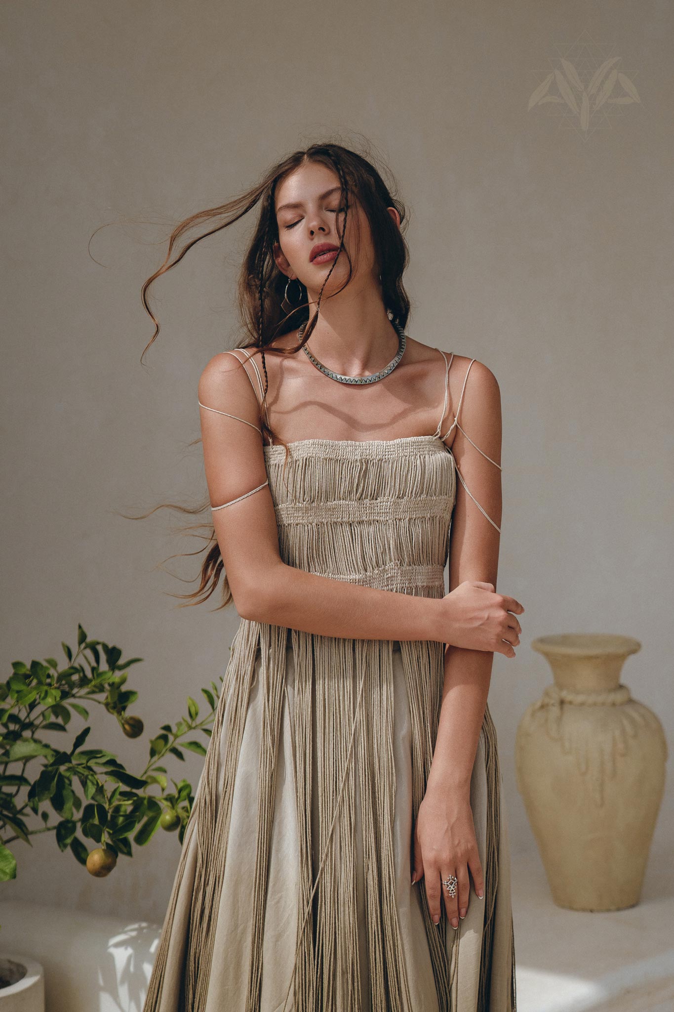 A woman in the Sage Green Tribal Goddess Slip Over by AYA Sacred Wear stands with eyes closed, long hair blowing, wearing delicate jewelry. Behind her, a green plant and large clay vase rest against a neutral wall.