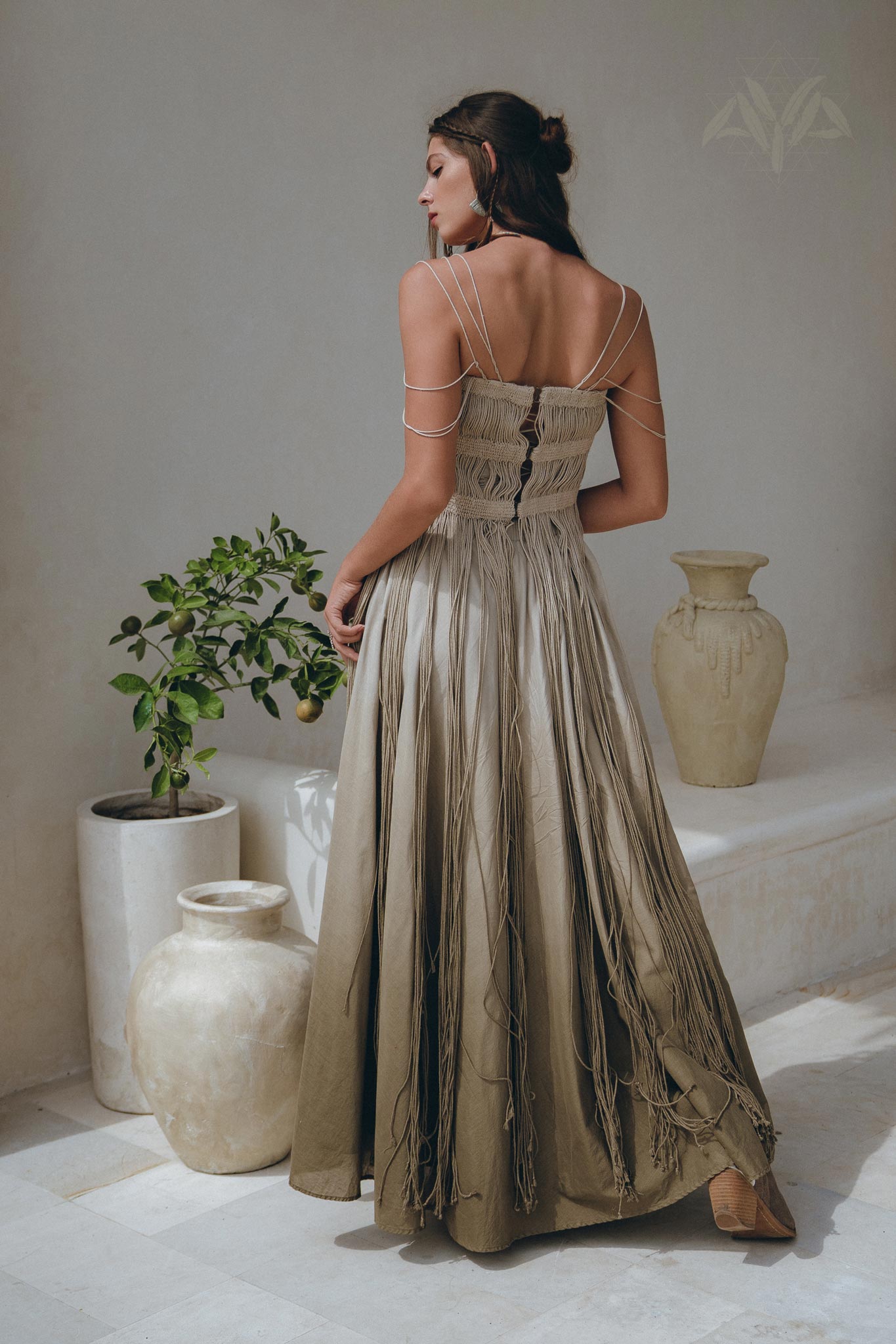 A woman in the Sage Green Tribal Goddess Slip Over by AYA Sacred Wear stands with her back turned, beside cream ceramic vases and a potted plant, all set against a light minimalist background.