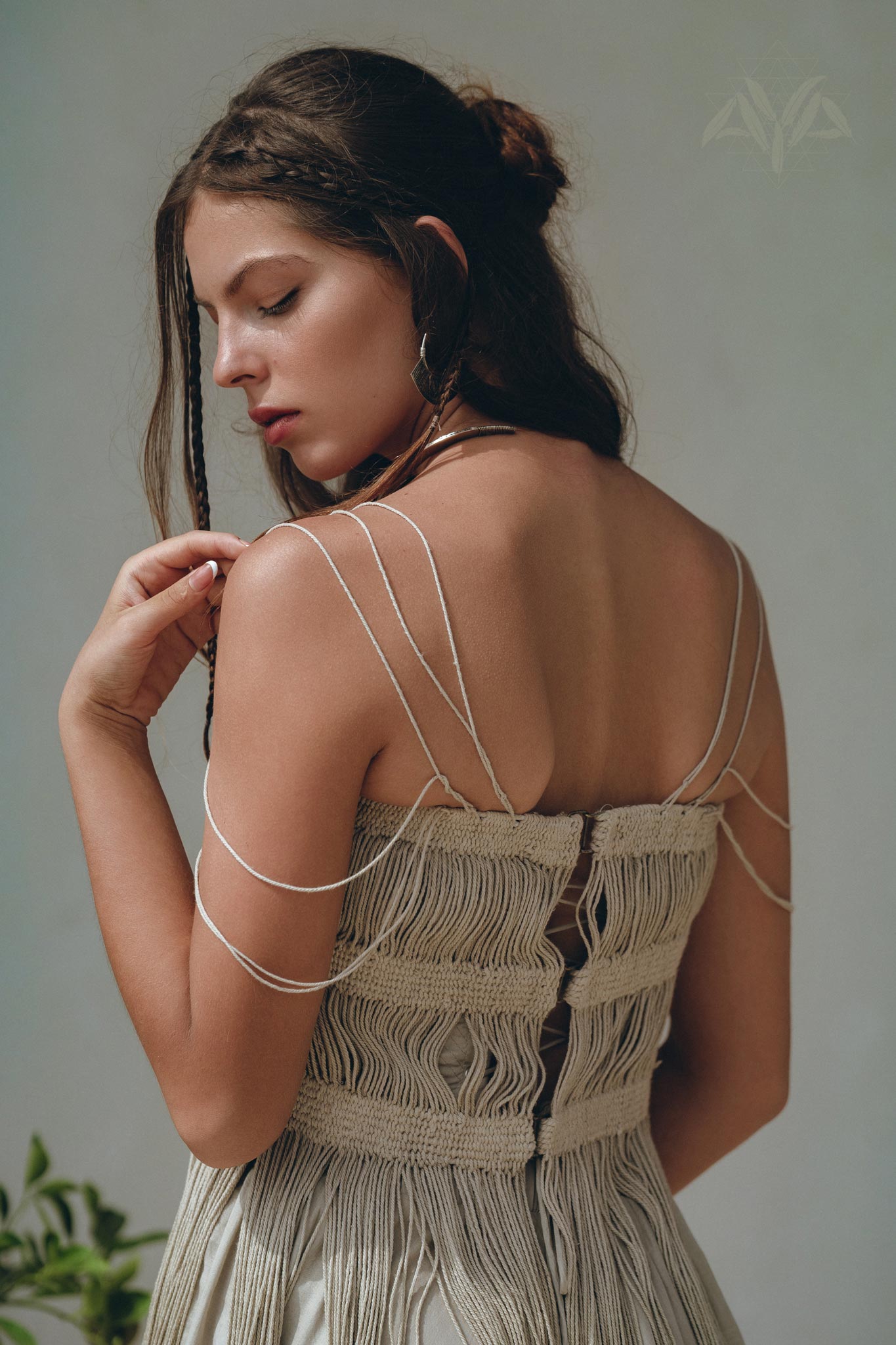 A woman with long brown hair in a braid wears the Sage Green Tribal Goddess Slip Over by AYA Sacred Wear—an ethnic, handwoven macrame sheer net dress—standing sideways and looking down thoughtfully.