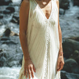A woman wearing a See Through Dress, a Long Maxi Dress by AYA Sacred Wear, stands by a rocky stream. The sunlight enhances the textured fabric of the Tribal Raw Cotton Cover-up and her long hair. The natural setting creates a serene and peaceful ambiance in the scene.