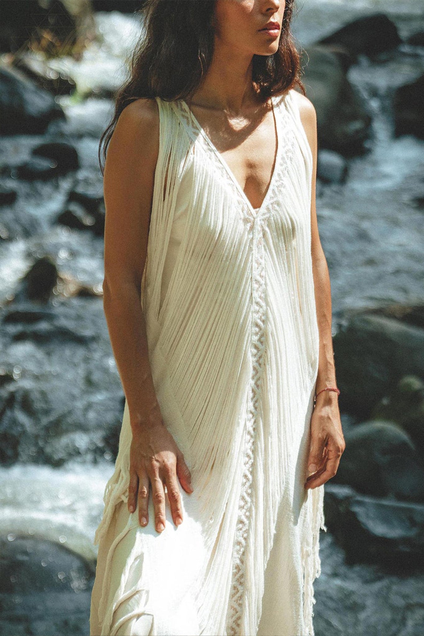 A woman wearing a See Through Dress, a Long Maxi Dress by AYA Sacred Wear, stands by a rocky stream. The sunlight enhances the textured fabric of the Tribal Raw Cotton Cover-up and her long hair. The natural setting creates a serene and peaceful ambiance in the scene.