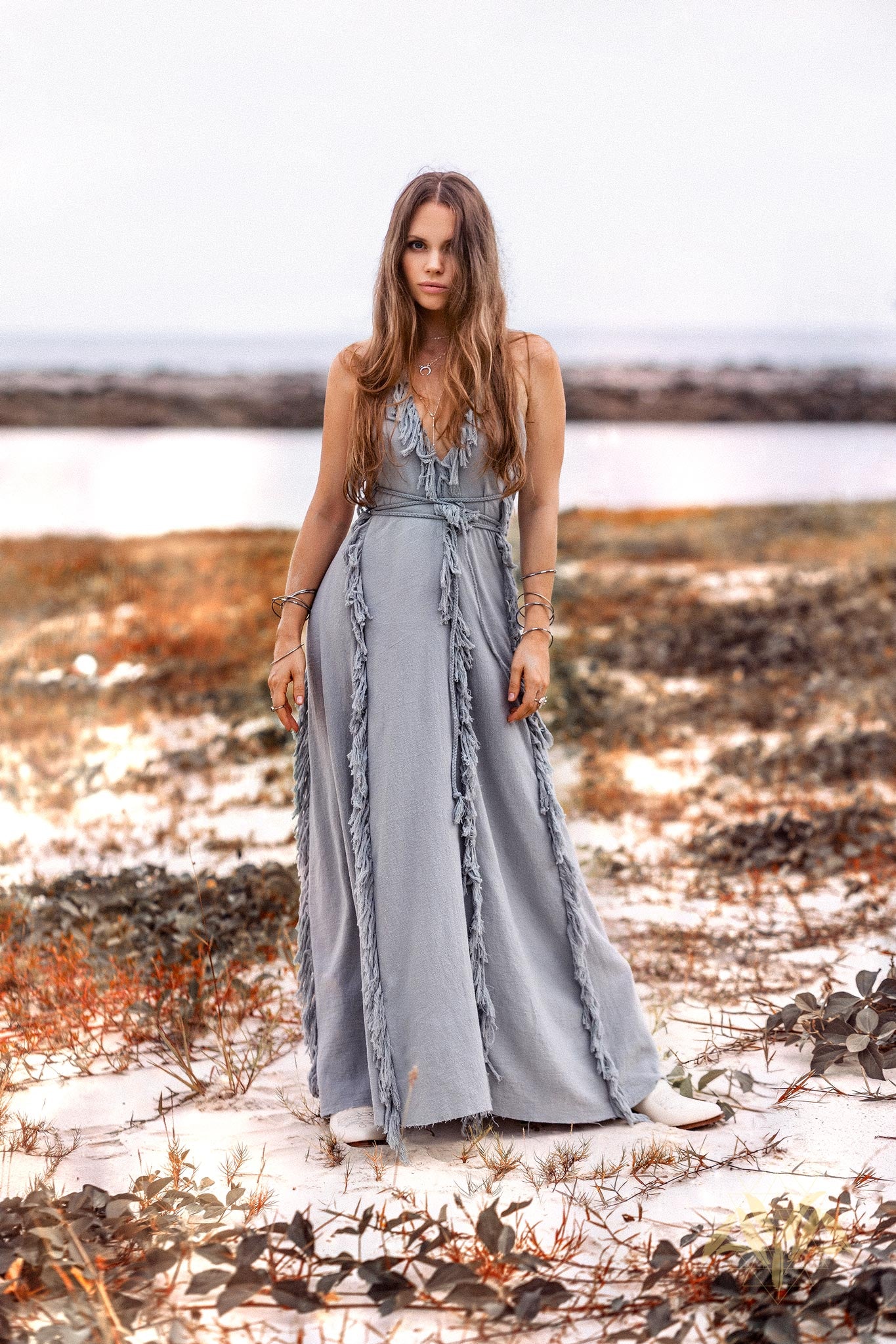 Full-length editorial shot of a model walking on a sandy beach wearing the Sky Blue raw cotton maxi dress with exquisite hand-loomed tassel fringe by AYA Sacred Wear.