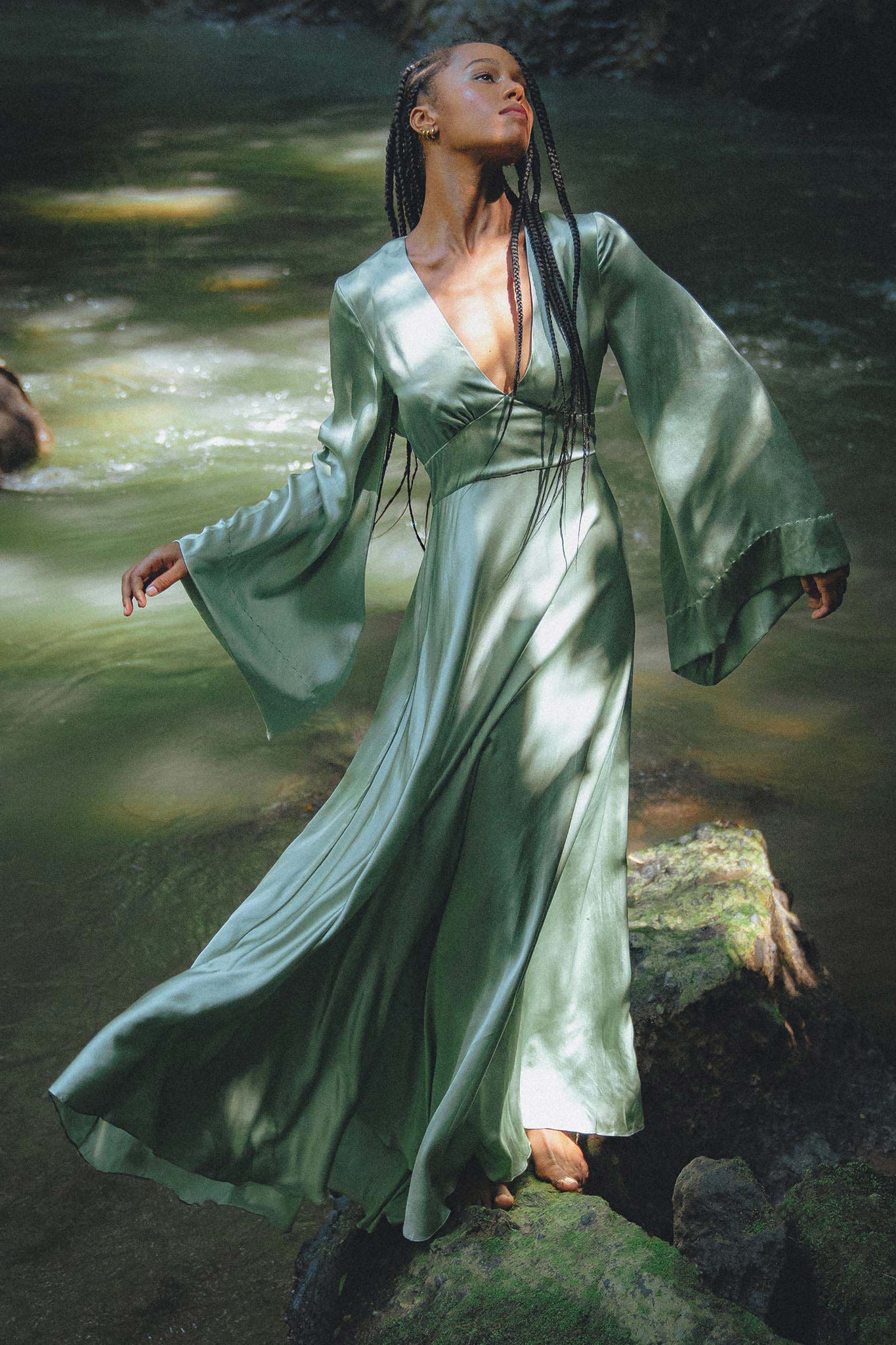 A woman with long braids is gracefully standing in a flowing dress with bell sleeves, surrounded by a serene natural setting. The soft fabric of the dress catches the sunlight, creating a magical effect. She is positioned on rocks near a gentle stream, embodying an ethereal vibe. This is the sky blue goddess dress, designed as a bridesmaid dress by Aya Sacred Wear.