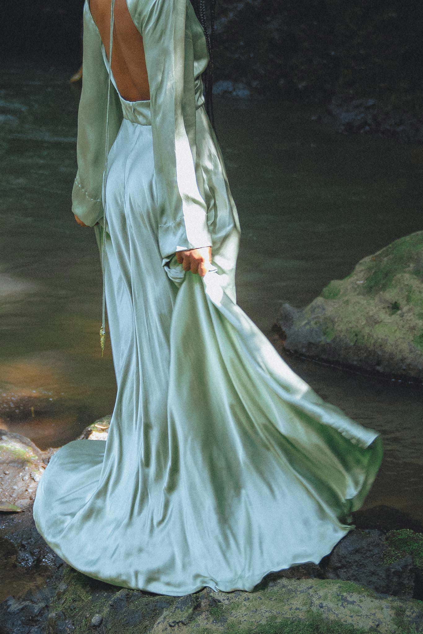 A model wearing a flowing, sky blue goddess dress from Aya Sacred Wear stands by a serene water setting, showcasing the graceful back and long sleeves of the garment as the fabric drapes elegantly around her. The dress catches the light, highlighting its silky texture and movement.