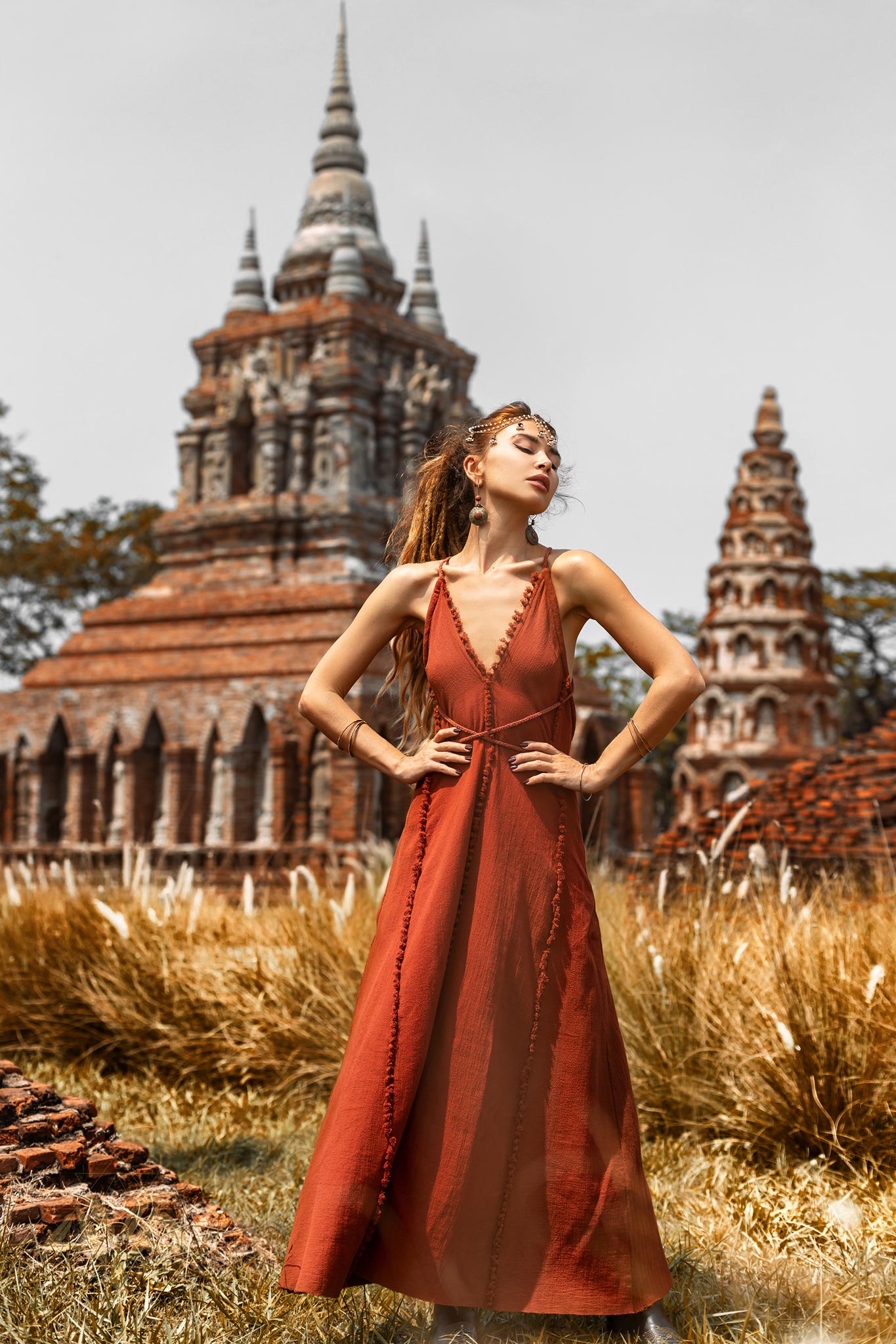 Wearing the handmade Goddess V Neck Dress by AYA Sacred Wear, a woman confidently stands with hands on hips in front of an ancient brick temple with ornate spires. Tall grass surrounds the area beneath a clear sky.