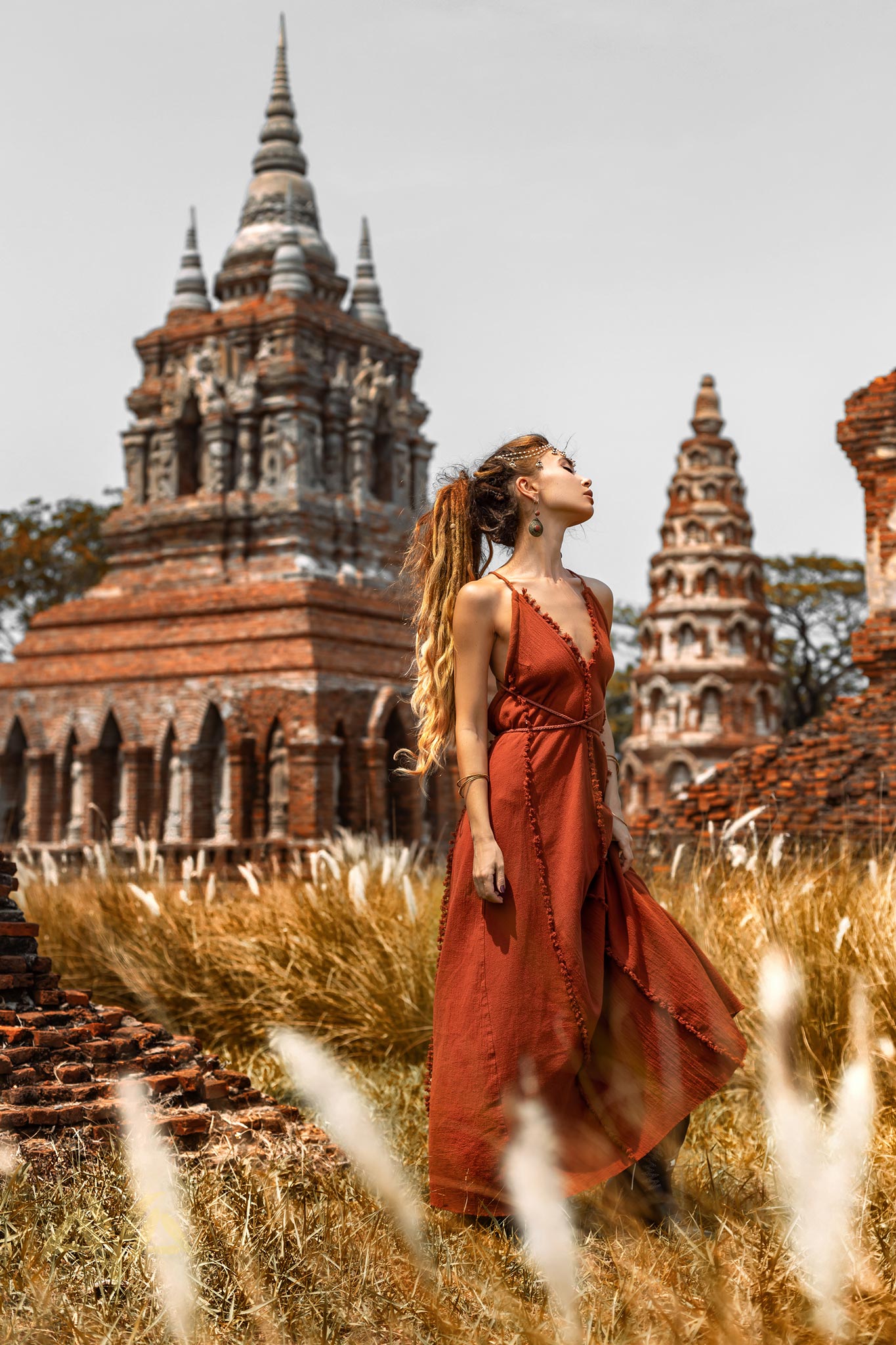 A woman in an AYA Sacred Wear Red Boho Bridesmaid Dress poses gracefully in a grassy area, with ancient brick pagodas behind her, exuding historical exploration and tranquility.