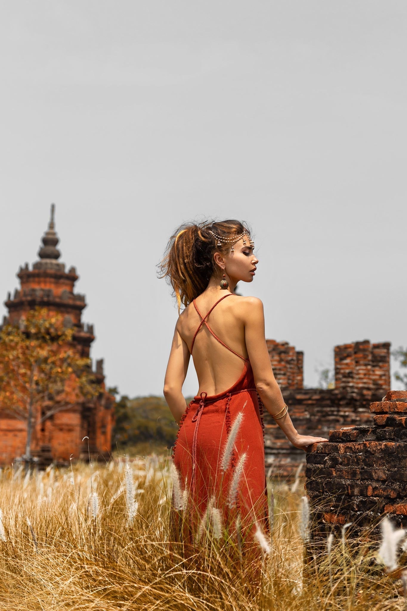Dressed in AYA Sacred Wear's Red Boho Bridesmaid Dress, a woman stands in a field of tall grass, gazing at an ancient brick structure with a pagoda-like tower. The overcast sky enhances the mysterious aura of this historical site, evoking another era.