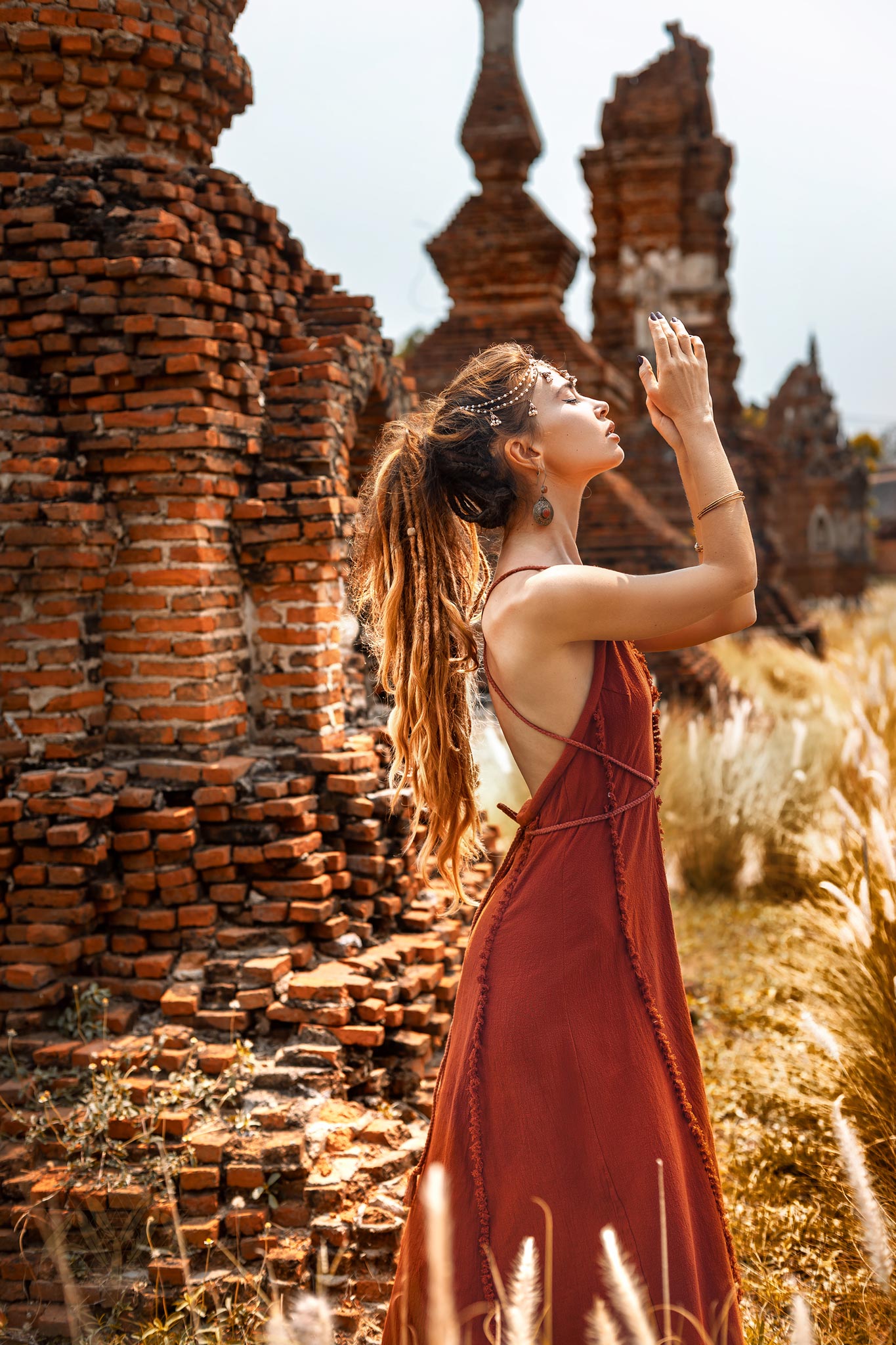 Wearing the Red Boho Bridesmaid Dress by AYA Sacred Wear, a woman stands among ancient brick ruins. The backless, flowing design complements her bohemian braids as she gazes upward with hands raised. The sun casts a warm glow, highlighting tall grasses in the background.