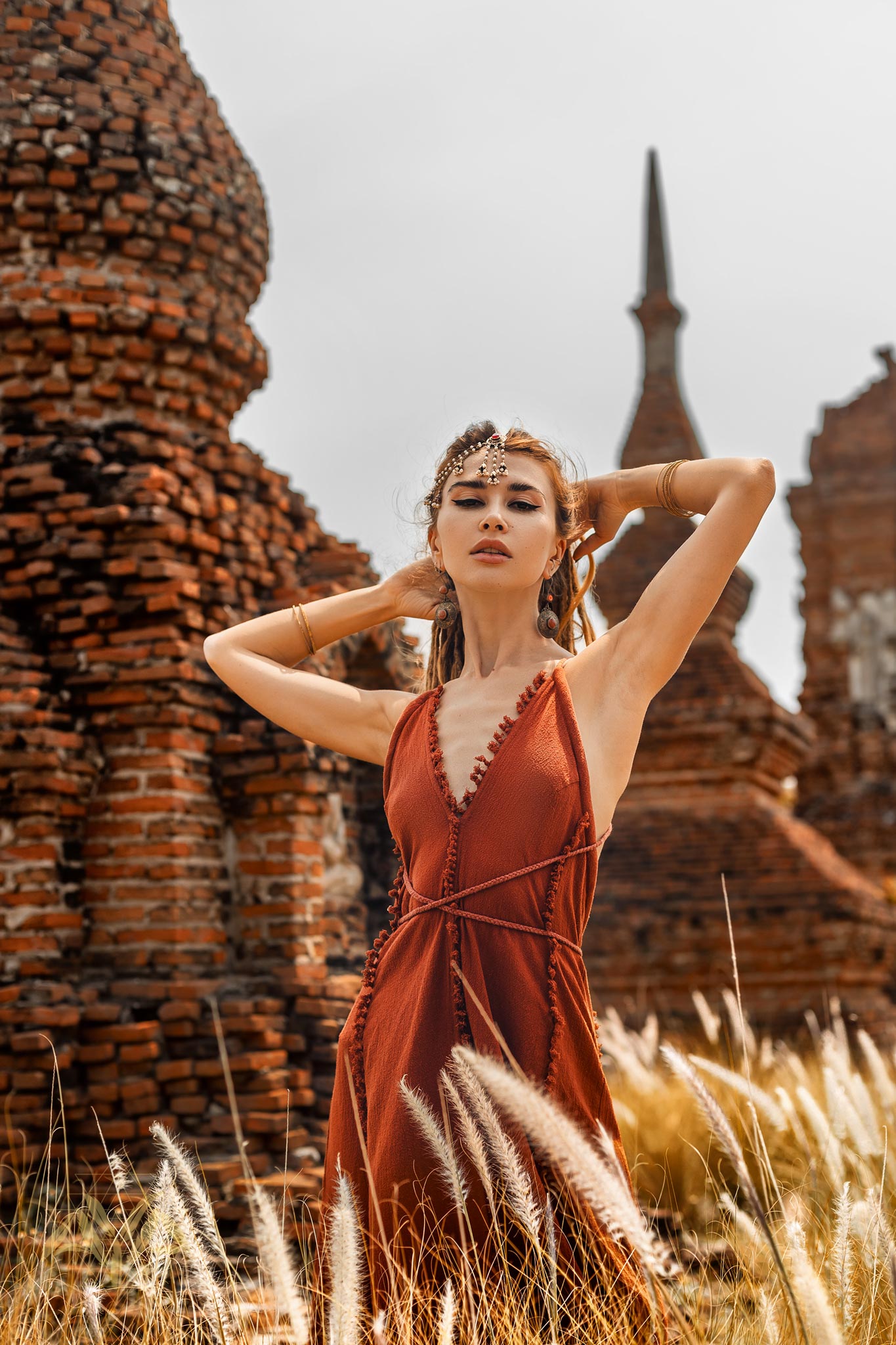 A woman poses against ancient brick structures in a Red Boho Bridesmaid Dress by AYA Sacred Wear, her hands behind her head, wearing a headpiece and bracelets. Standing amidst tall grass under a cloudy sky, she exudes an effortlessly chic vibe.