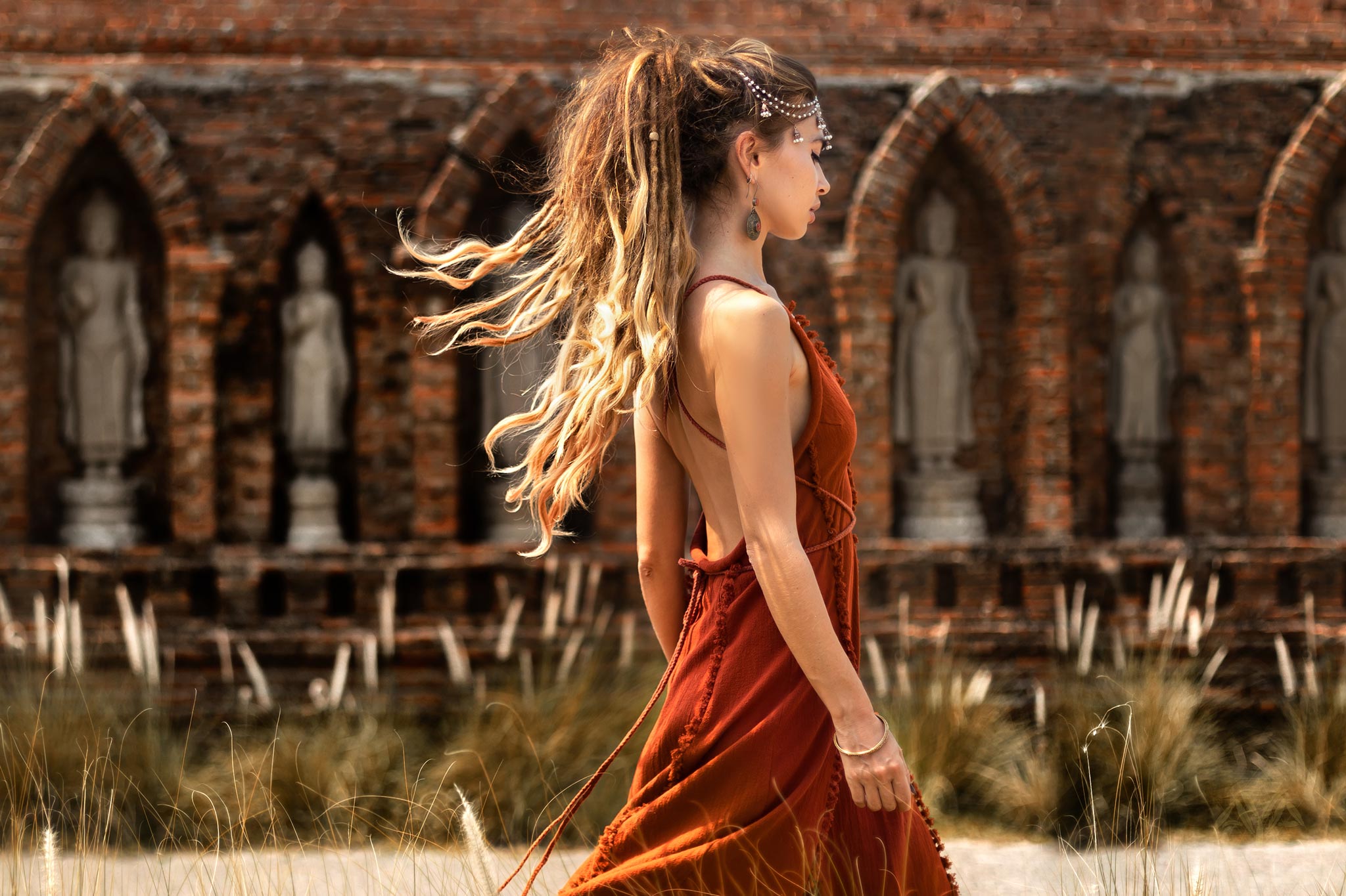 A woman with long hair in a ponytail, wearing an AYA Sacred Wear Red Boho Bridesmaid Dress, walks past brick arches and statues. Tall grass sways gently in the foreground, adding movement and tranquility to the scene.