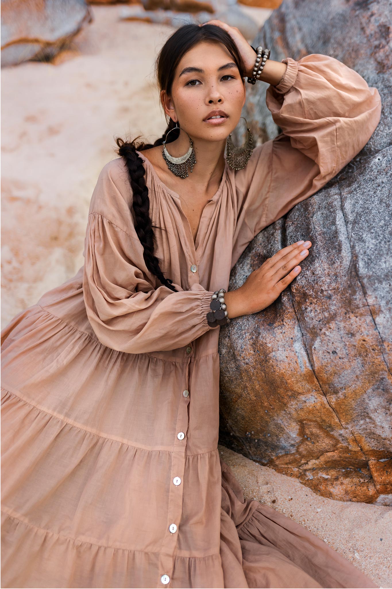 Against a rocky backdrop, a person with long braided hair poses outdoors wearing AYA Sacred Wear's Boho Dress for Women in dusty pink, paired with large circular earrings. The soft cotton fabric and natural surroundings create a serene, bohemian ambiance.