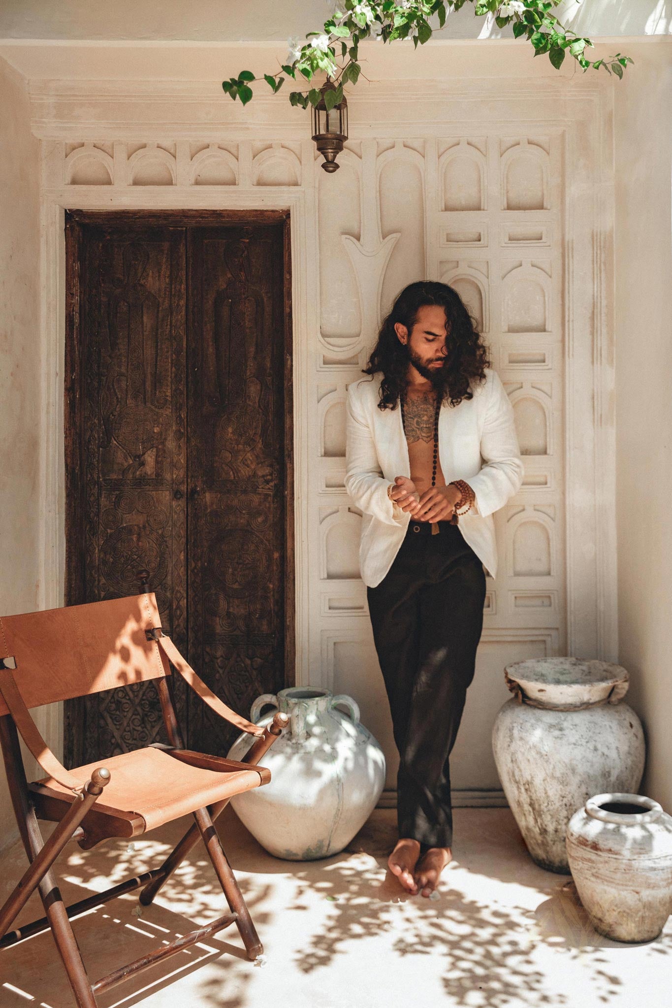 Male model with long hair wearing an off-white fine linen boho blazer and dark pants, leaning against a rustic carved wooden door.