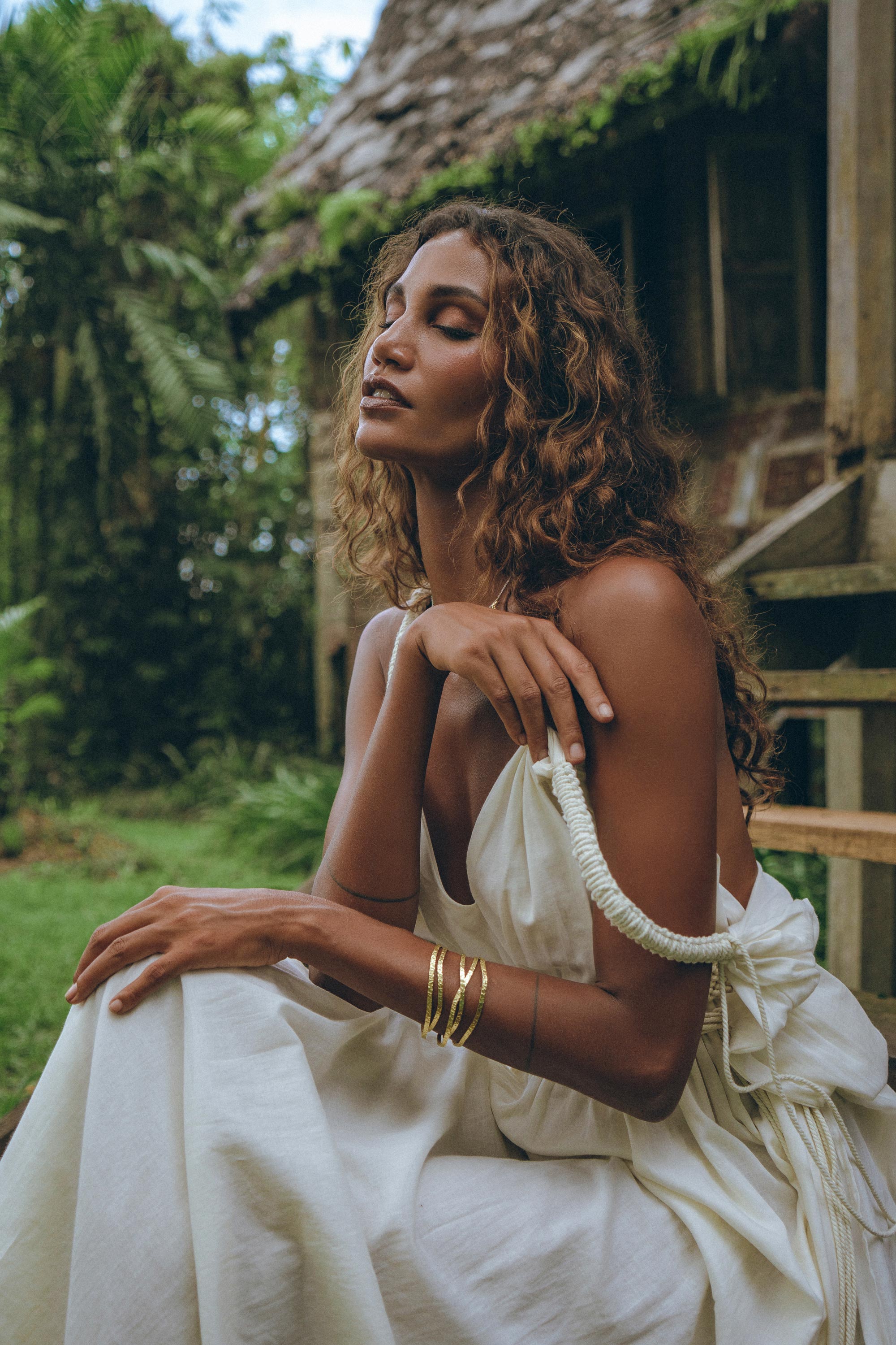 A woman with curly hair sits outside near a rustic wooden structure, wearing the Boho Beach Macrame Wedding Dress by AYA Sacred Wear. This off-white, multiway bohemian dress complements her relaxed demeanor as she closes her eyes. Gold bracelets adorn her wrists amid the lush green foliage that surrounds the area.