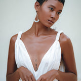 A person with short hair is seen standing against a neutral background, wearing the Boho Beach Macrame Wedding Dress by AYA Sacred Wear. The off-white multiway bohemian dress showcases their geometric chest tattoos as they gaze to the side, with one hand clasping the other. An artisanal macrame belted waist completes this unique look.