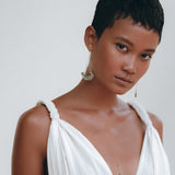 A person with short hair is standing against a plain background, adorned in the Boho Beach Macrame Wedding Dress by AYA Sacred Wear. The dress, an off-white multiway bohemian creation, is made of light homespun cotton fabric. They have a tattoo on their chest and are wearing large earrings, exuding a calm and composed expression.