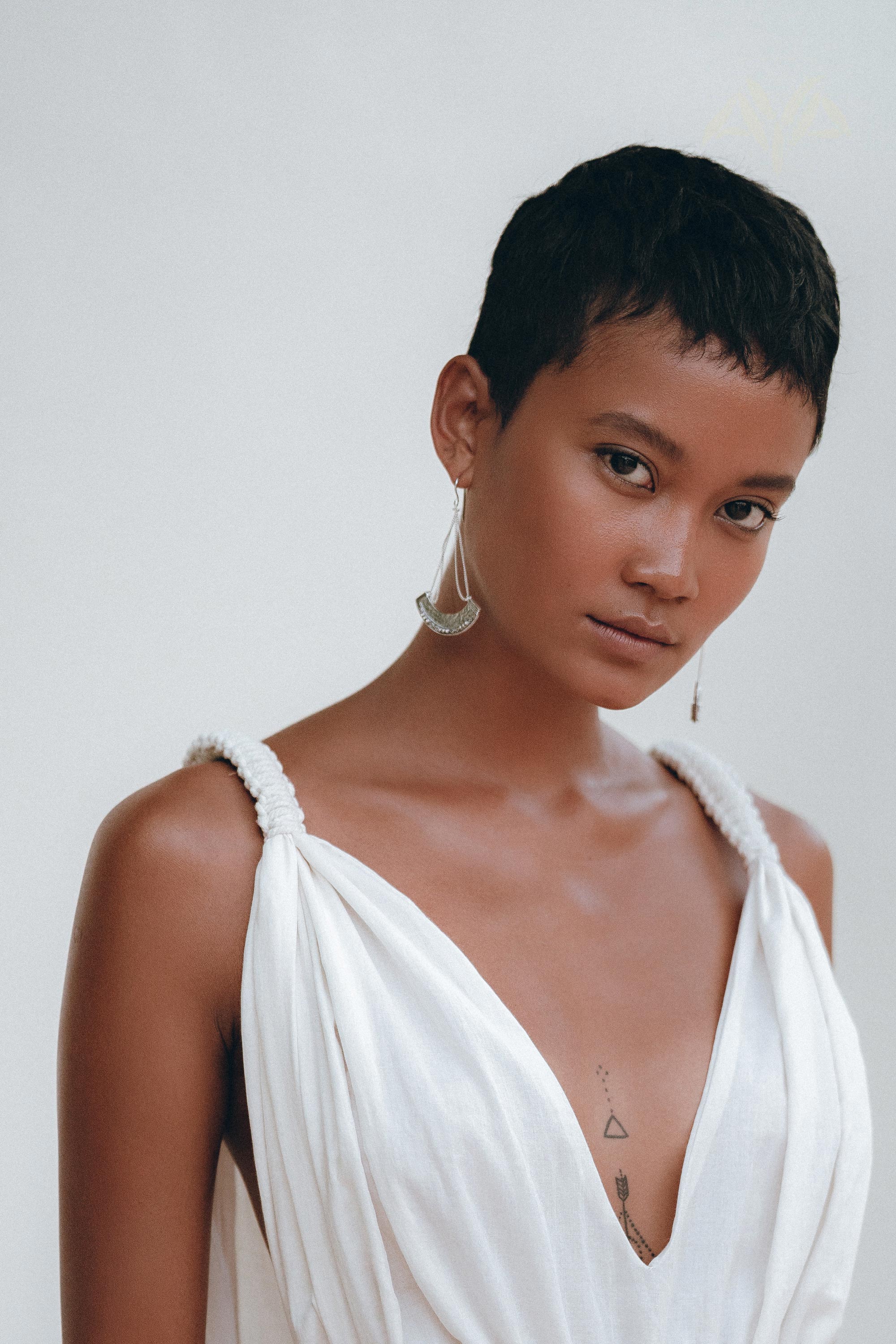 A person with short hair is standing against a plain background, adorned in the Boho Beach Macrame Wedding Dress by AYA Sacred Wear. The dress, an off-white multiway bohemian creation, is made of light homespun cotton fabric. They have a tattoo on their chest and are wearing large earrings, exuding a calm and composed expression.
