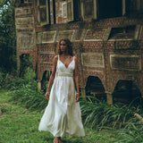 A woman walks barefoot on lush grass beside an intricately carved, rustic wooden structure with open windows, wearing an off-white multiway bohemian dress by AYA Sacred Wear. The setting is tropical and surrounded by greenery, evoking a serene and natural atmosphere that embodies the essence of the Boho Beach Macrame Wedding Dress.