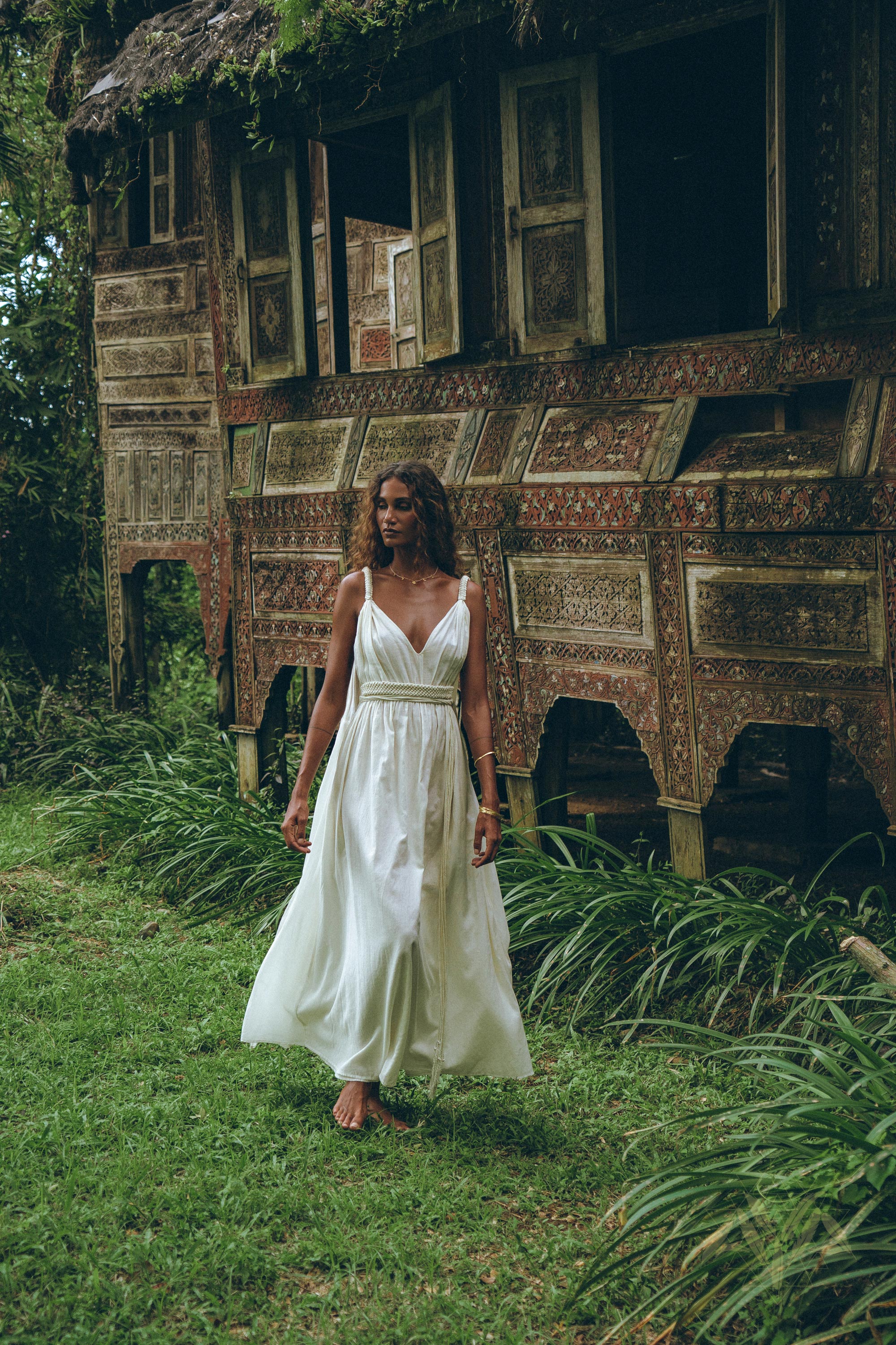 A woman walks barefoot on lush grass beside an intricately carved, rustic wooden structure with open windows, wearing an off-white multiway bohemian dress by AYA Sacred Wear. The setting is tropical and surrounded by greenery, evoking a serene and natural atmosphere that embodies the essence of the Boho Beach Macrame Wedding Dress.