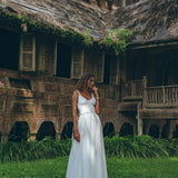 Dressed in an AYA Sacred Wear Boho Beach Macrame Wedding Dress, an Off-White Multiway Bohemian piece, a woman gazes thoughtfully in front of a traditional wooden house adorned with intricate carvings and lush greenery. This aged structure, resembling a unique piece of art, features a weathered roof and decorative patterns on its walls.