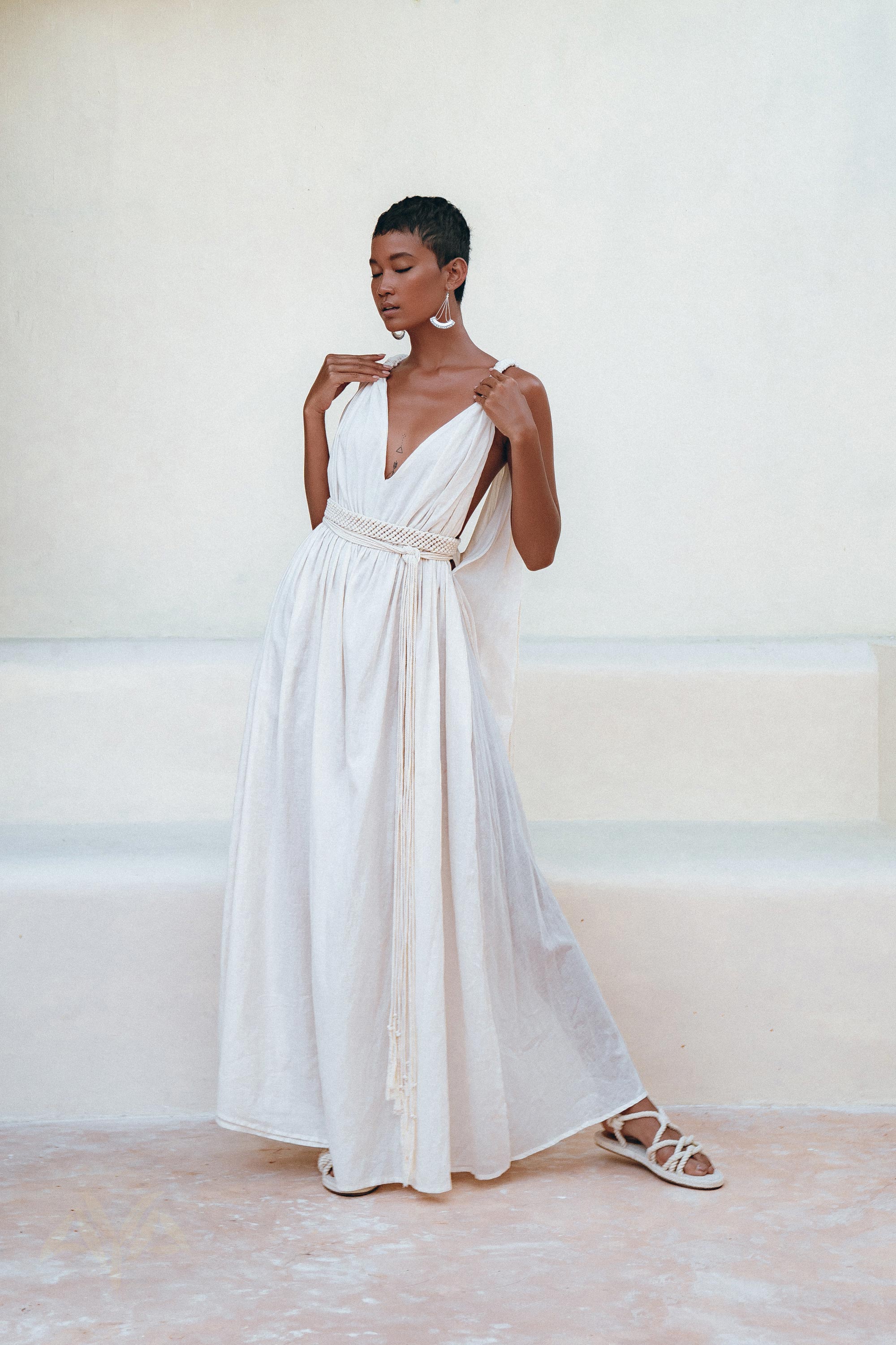 Against a minimalist backdrop, a person poses gracefully in the Boho Beach Macrame Wedding Dress by AYA Sacred Wear. This off-white, multiway bohemian dress features a flowing, sleeveless design crafted from light homespun cotton fabric with a deep V-neckline and cinched waist. Paired with white sandals and large hoop earrings, they exude the enchanting charm of this boho-inspired attire.