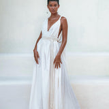 Against a light background, a confident individual with short hair showcases the elegance of the AYA Sacred Wear's Boho Beach Macrame Wedding Dress. Featuring a deep V-neck and tied waist, this off-white multiway bohemian dress is crafted with homespun cotton, perfectly accessorized with hoop earrings, macrame belted details, and white sandals.