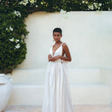 A person in an off-white multiway bohemian dress by AYA Sacred Wear stands gracefully against a pale wall, surrounded by lush greenery and white flowers. With short hair and strappy sandals, they exude elegance in a serene outdoor setting, their attire enhanced by the macrame belted details of the Boho Beach Macrame Wedding Dress.