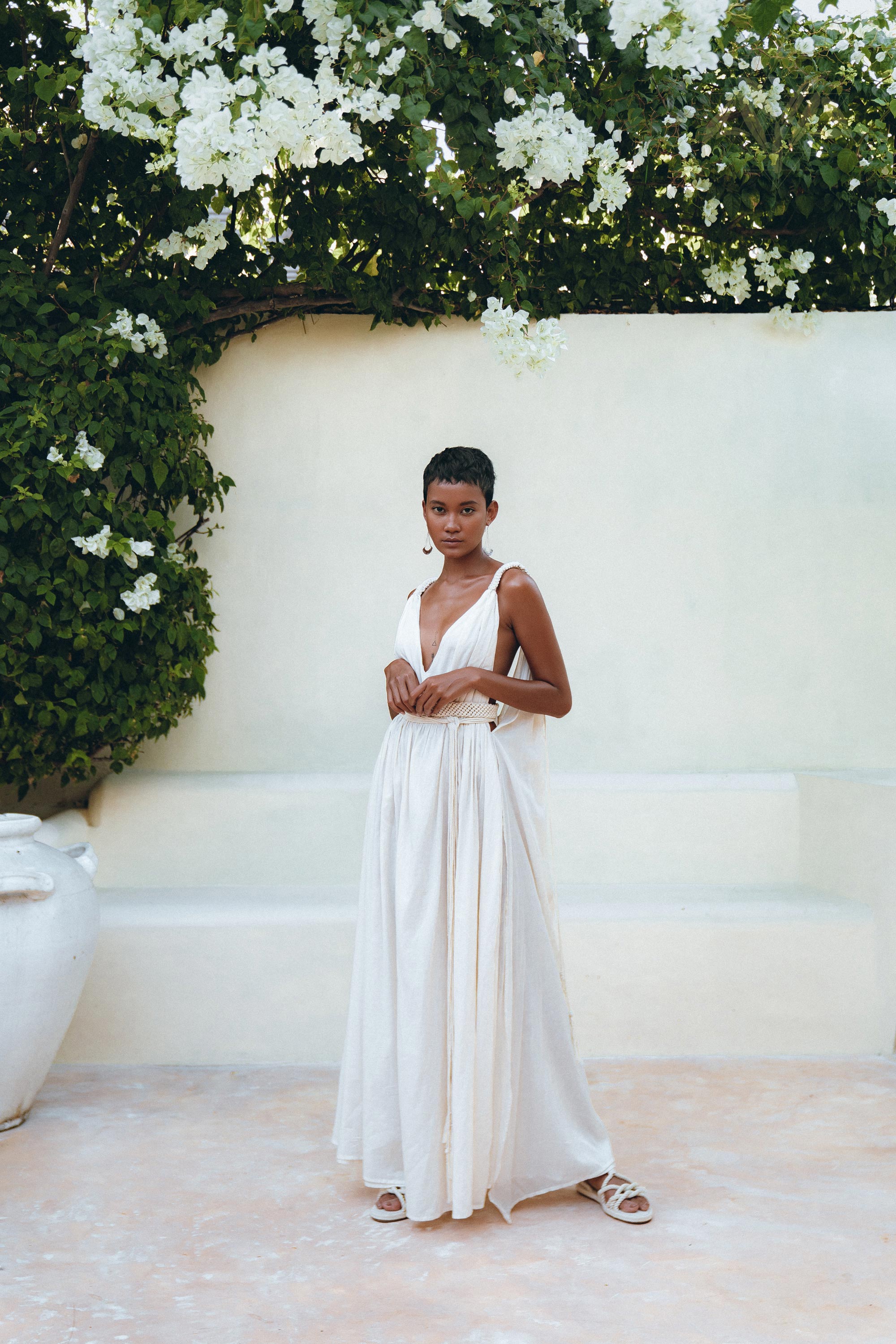 A person in an off-white multiway bohemian dress by AYA Sacred Wear stands gracefully against a pale wall, surrounded by lush greenery and white flowers. With short hair and strappy sandals, they exude elegance in a serene outdoor setting, their attire enhanced by the macrame belted details of the Boho Beach Macrame Wedding Dress.
