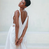 Wearing the Boho Beach Macrame Wedding Dress by AYA Sacred Wear, featuring an off-white multiway design, a person stands gracefully with their head tilted and hands clasped behind. The elegant bohemian dress beautifully complements the neutral outdoor backdrop, creating a scene reminiscent of a living piece of art.