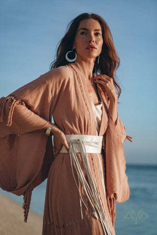 A woman stands on a beach, adorned in a flowing light pink garment, accessorized with an elegant white leather belt from AYA Sacred Wear. Made from pure organic genuine sheep leather, the belt adds a bohemian touch to her ensemble with its long tassels. Her loose hair complements her large earrings as the clear sky and ocean form a serene backdrop.